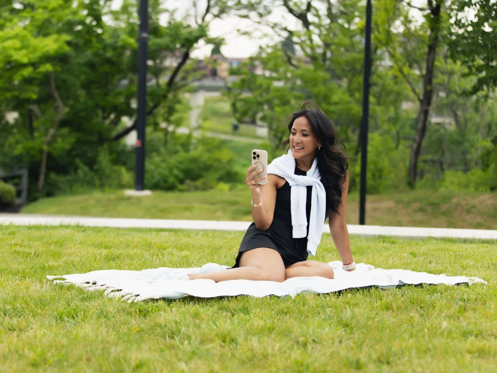 Woman on blanket in park, holding phone, smiling. Wearing black dress and white sweater.