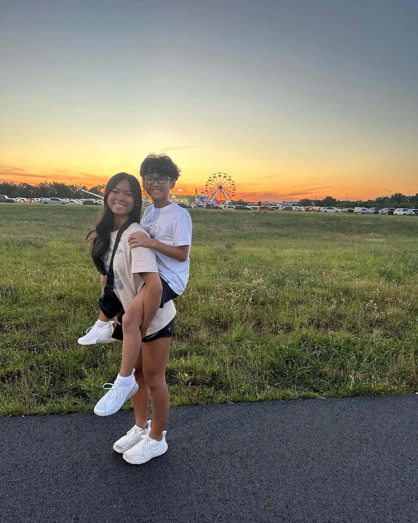 Woman giving piggyback ride to person with glasses, sunset and Ferris wheel in background.