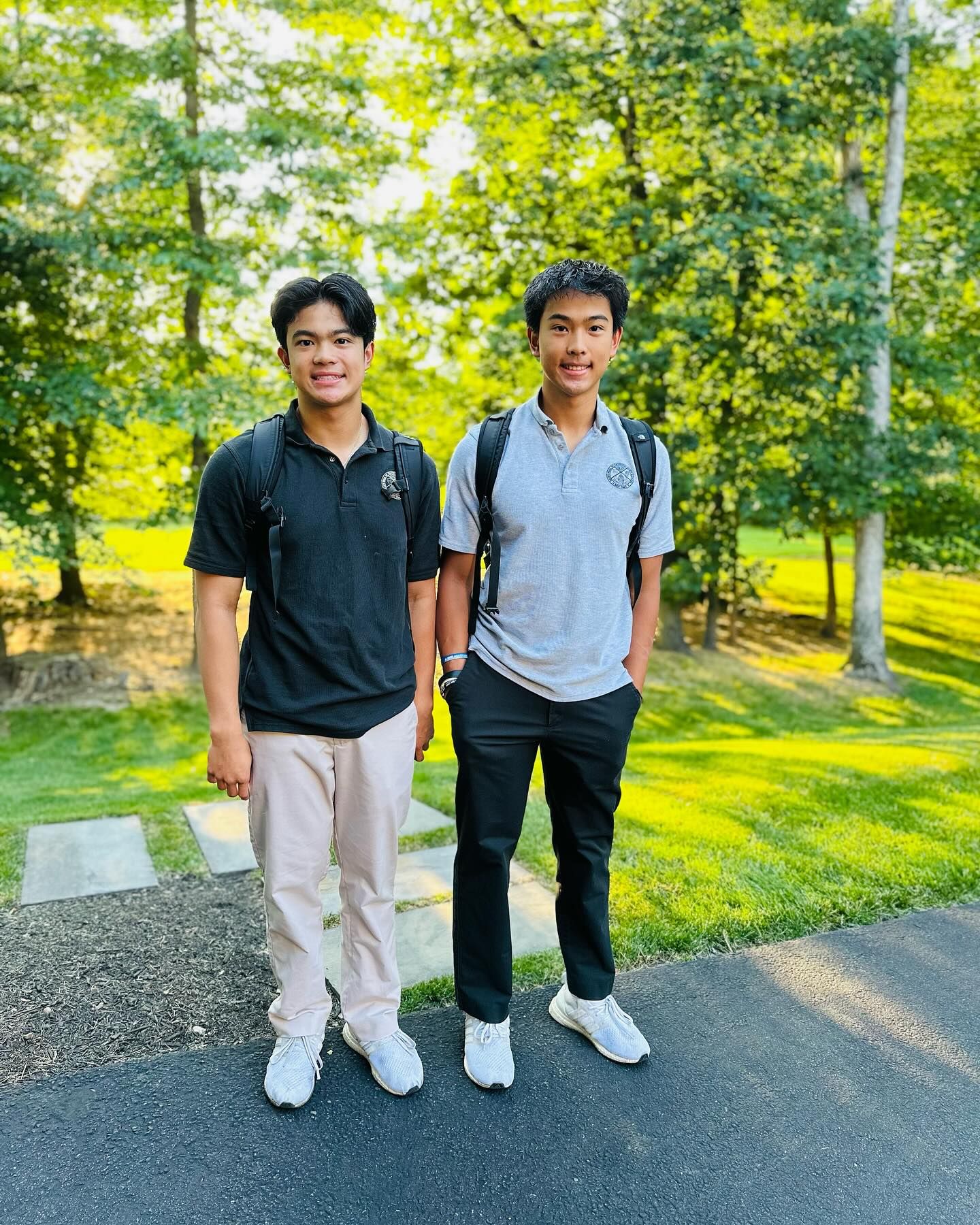 Two young men stand outside. One wears a black polo shirt and khaki pants, the other a grey polo shirt and black pants.