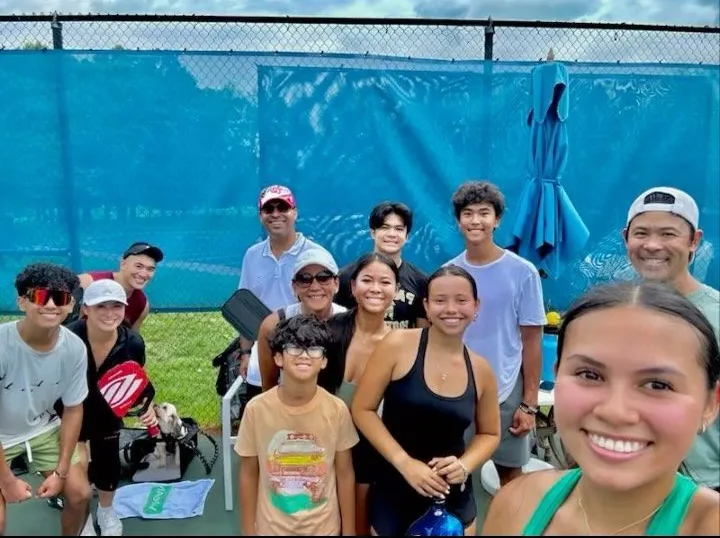 Group of people smiling at camera near a blue tennis court fence. Some hold paddles. Overcast sky.