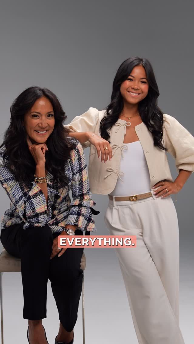Two women pose, one seated and one standing. They smile. Plain background.