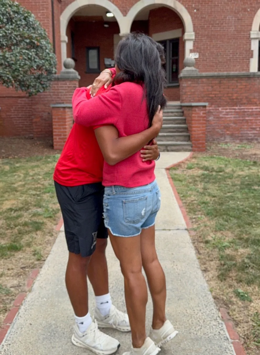 Two people embracing on a walkway in front of a brick building. One wears red, the other, denim shorts.