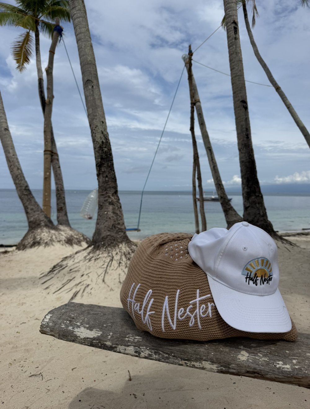 A Half Nester tote bag and cap resting on driftwood on a palm-lined tropical beach