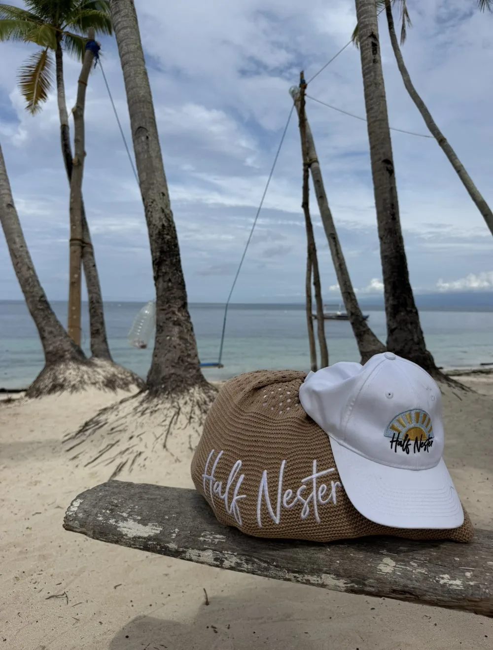 A Half Nester tote bag and cap resting on driftwood on a palm-lined tropical beach